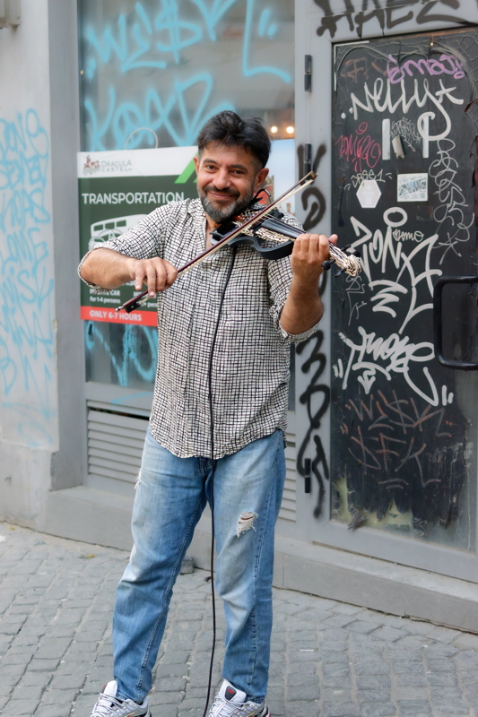 Person playing an electric violin against a graffitied wall, styled with a checkered button-down shirt and distressed blue jeans